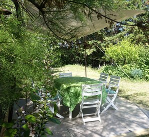 Une terrasse extérieure avec une table ronde et des chaises blanches, entourée d’une végétation luxuriante et protégée du soleil par un grand parasol.