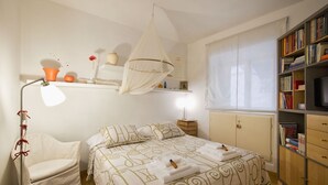 Quarto com cama, mesa de cabeceira, cadeira, estante e janela com cortinas.