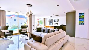Ein modernes Wohnzimmer mit einem großen Sofa, zwei Sesseln, einem Glastisch und einem Essbereich im Hintergrund.