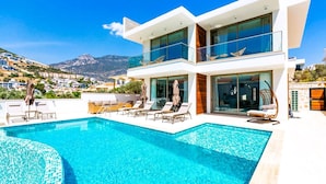 Ein modernes, zweistöckiges Gebäude mit Pool auf dem Dach, Liegestühlen und Blick auf die umliegende Landschaft.
