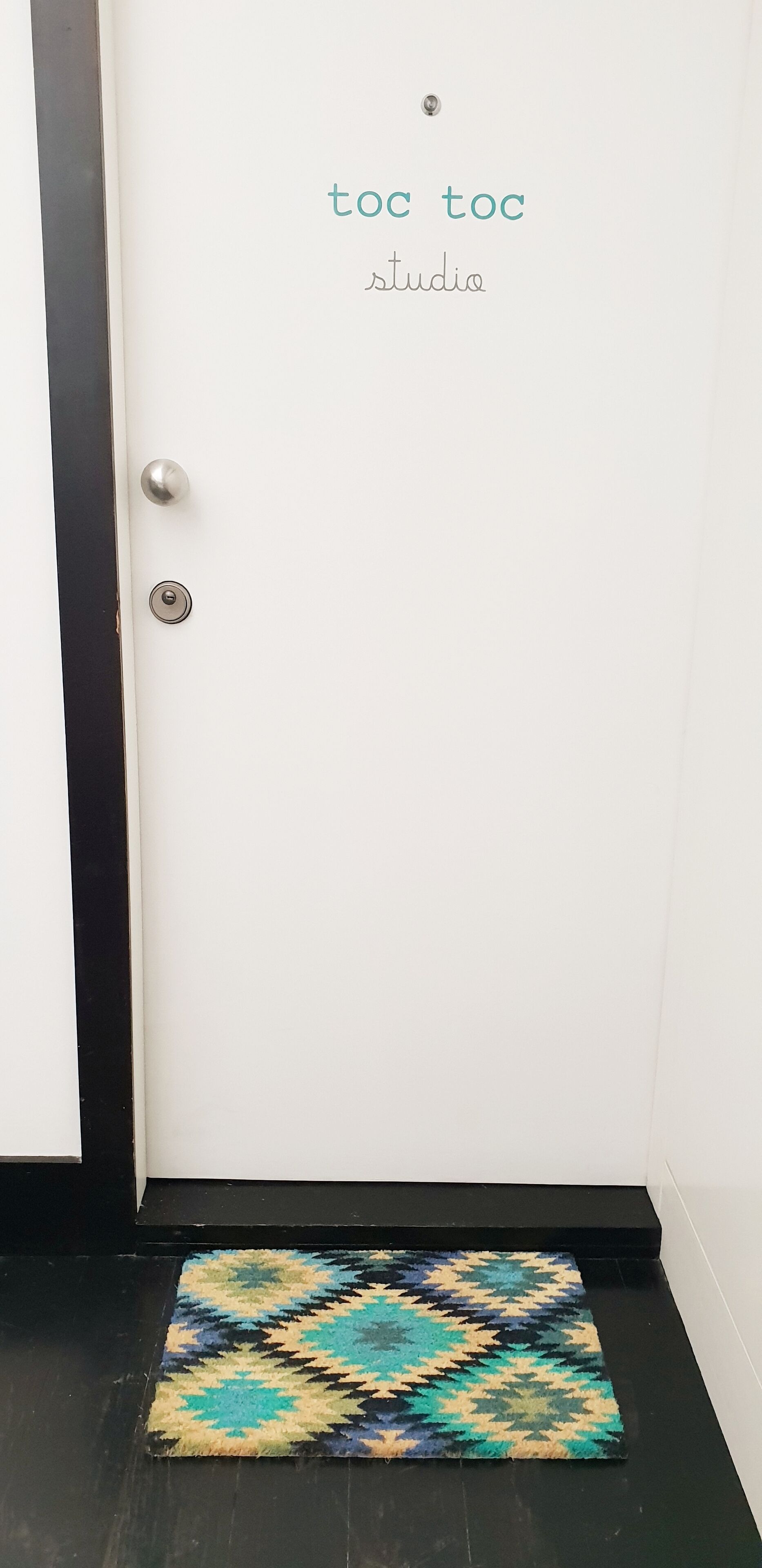 Een witte deur met de tekst 'toc toc studio', een metalen handvat en een gekleurd tapijt bij de ingang.