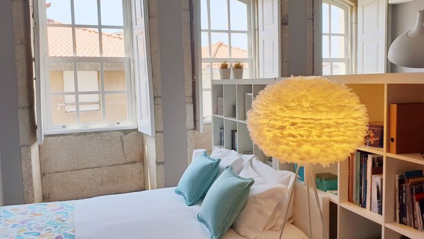 Een slaapkamer met een bed, kussens, een boekenkast en een raam met gordijnen.