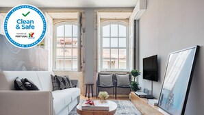 Een moderne woonkamer met een witte bank, een bijzettafeltje met een boek en bloemen, twee zwarte stoelen, een televisie en een groot raam met uitzicht op gebouwen.