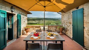 An outdoor patio with a table set for two, surrounded by stone walls and green shutters, overlooking a scenic landscape.