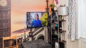 A room with a large flat-screen TV mounted on the wall, a bookshelf with various items, a clock, and a decorative lamp.