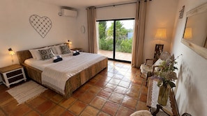 A bedroom with a large bed, a nightstand, a chair, a vase with flowers, and a view of greenery outside. - Airconditioned villa 2 to 10 people, private swimming pool and lovely ocean view (benagil)