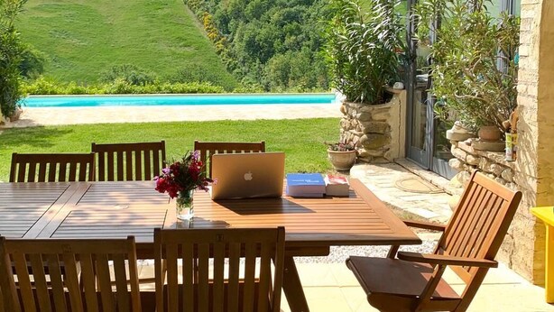 Une terrasse extérieure avec une table et des chaises en bois, donnant sur une piscine et des collines verdoyantes.