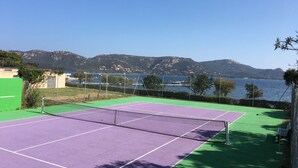 A tennis court with a net, surrounded by a green fence, with a view of a lake and mountains in the background. - Cosy, quiet villa 150 metres from the sea and swimming pool Tennis included (Porto-Vecchio)