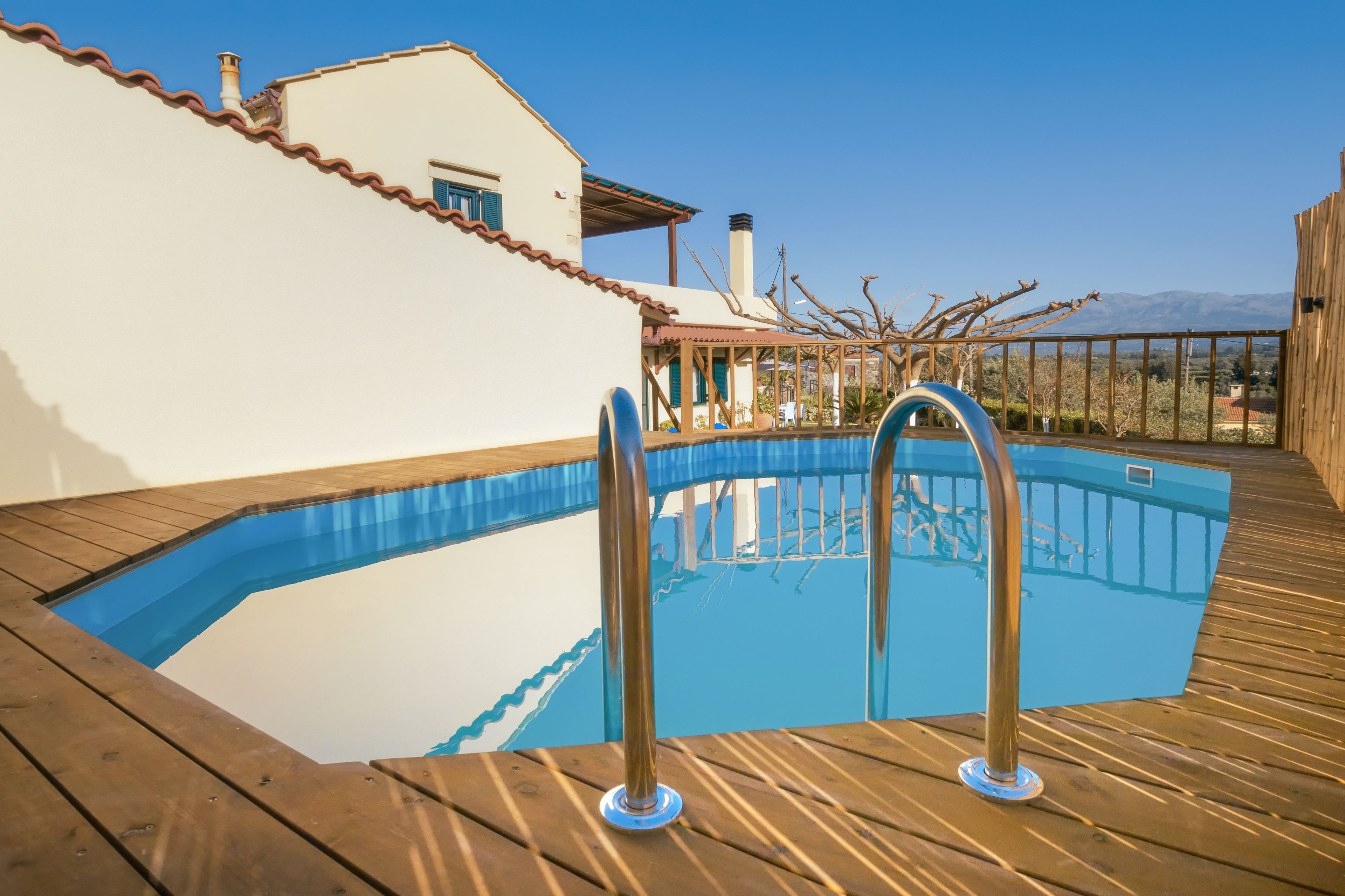 A swimming pool with a wooden deck, a white building with a red roof, and a clear blue sky in the background.
