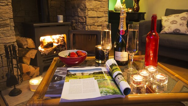 A cozy living room with a fireplace, a glass table with a magazine, candles, and bottles of wine.