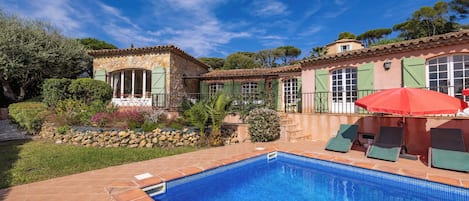 A swimming pool with a tiled edge, a red umbrella, and a stone building with green shutters.