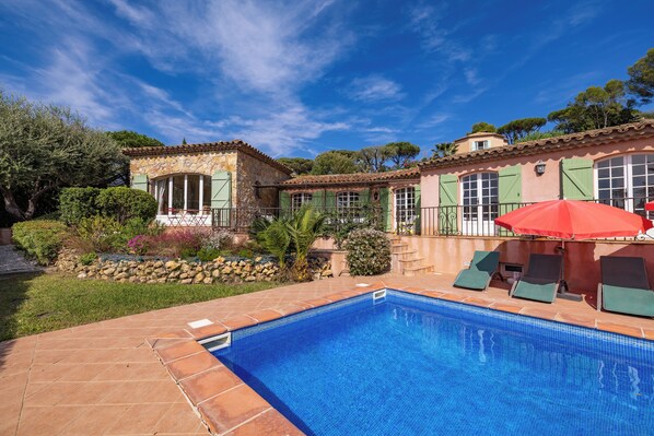 A swimming pool with a tiled edge, a red umbrella, and a stone building with green shutters.