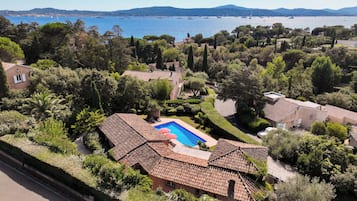 Un quartier résidentiel doté d’une piscine, entouré de bâtiments aux toits de tuiles en terre cuite, d’une végétation luxuriante et offrant une vue sur la mer et les bateaux.