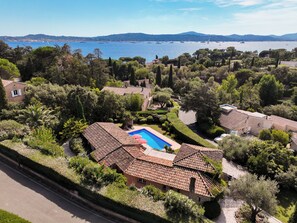 A residential area with a swimming pool surrounded by terracotta-tiled buildings, lush greenery, and a view of the sea with boats.