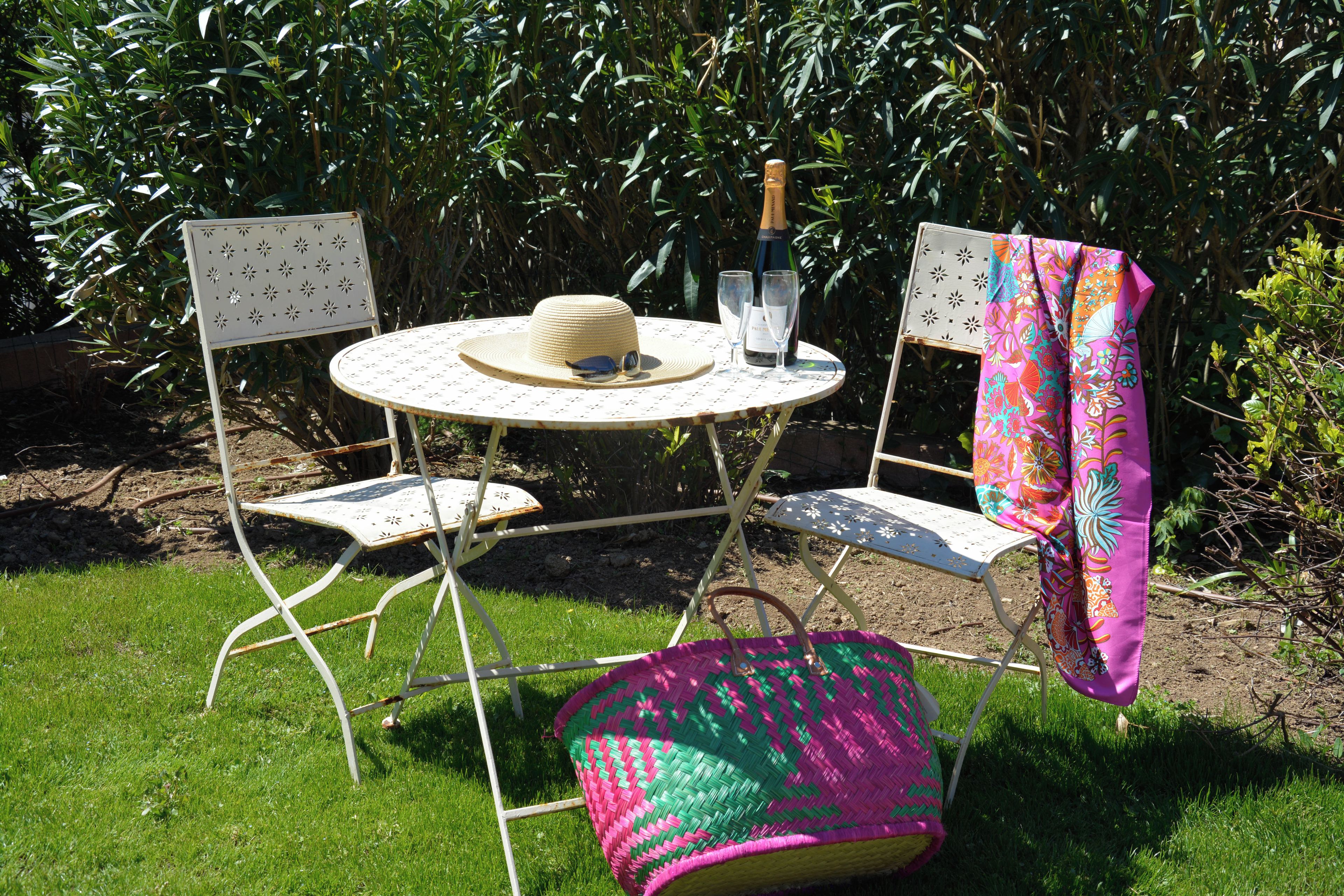 A round metal table with a hat and wine glasses on it, surrounded by two metal chairs with a colorful cloth draped over one.