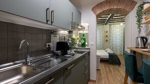 Eine kompakte Küche mit modernem Waschbecken, eingebauten Schränken und einer Kaffeemaschine. Ein kleiner Essbereich mit Tisch und Stühlen ist sichtbar, und ein Ziegelbogen führt in ein Schlafzimmer mit Bett und Nachttisch.