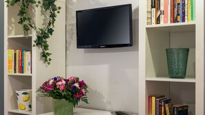 Ein Zimmer mit einem wandmontierten Flachbildfernseher, einem Bücherregal mit Büchern, einer Vase mit Blumen und einer Fernbedienung auf einem kleinen Tisch.