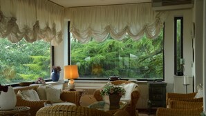 Una habitación acogedora con muebles de mimbre, un ventanal con cortinas, un jarrón con flores y una lámpara.