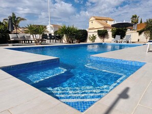 Una piscina con bordes de azulejos, rodeada de sillones y sombrillas, con edificios al fondo.