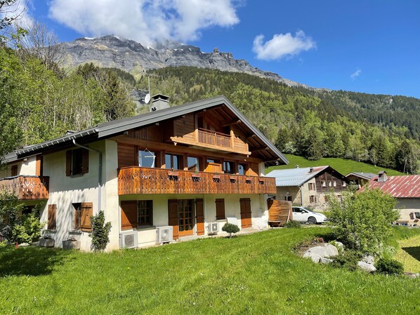 A two-story wooden chalet with balconies, surrounded by lush greenery and mountains in the background.