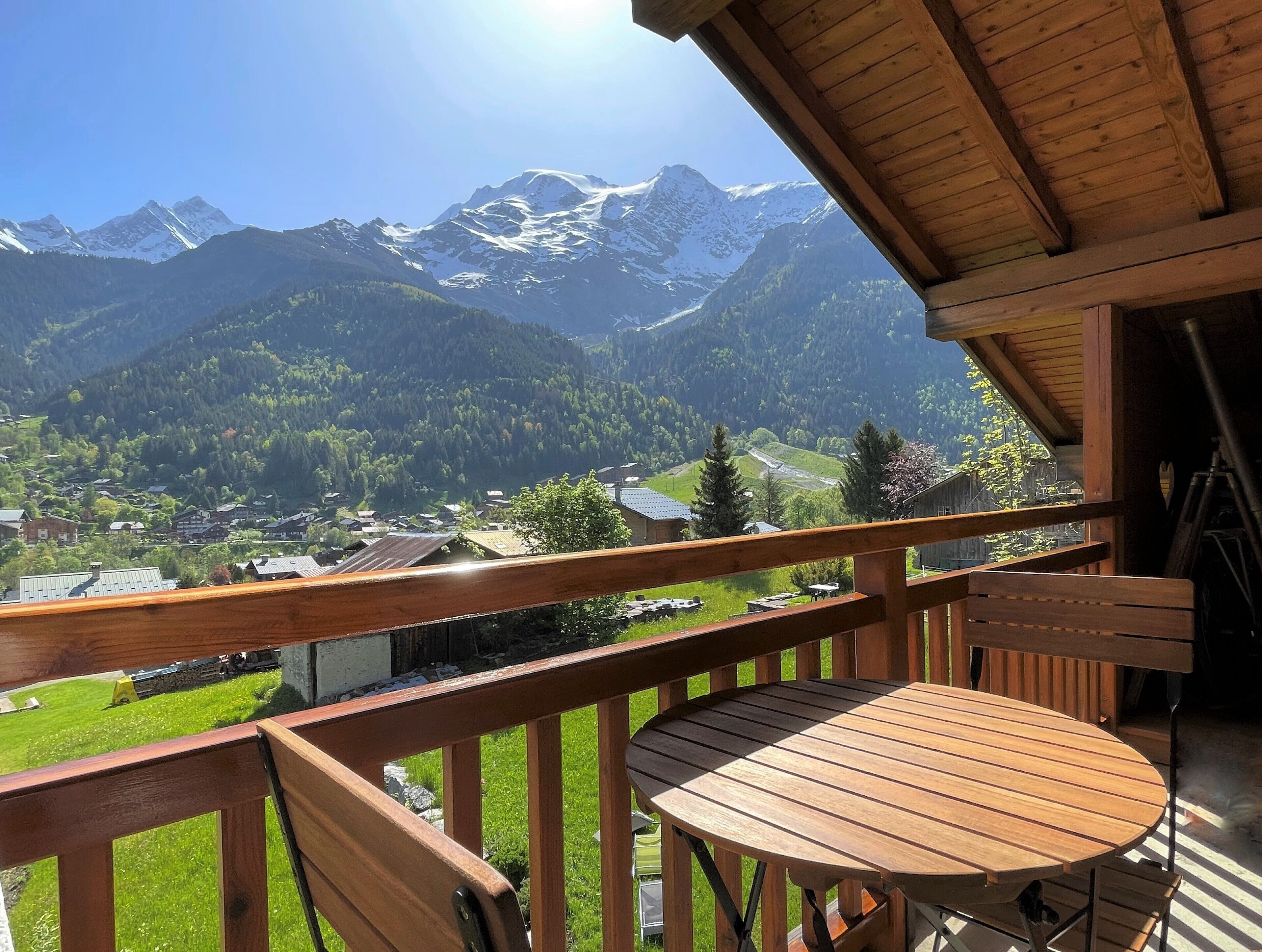 Un balcon en bois, agrémenté d’une table et de chaises, qui offre une vue sur un paysage montagneux avec des sommets enneigés et un village en contrebas.