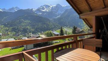 Un balcon en bois, agrémenté d’une table et de chaises, qui offre une vue sur un paysage montagneux avec des sommets enneigés et un village en contrebas.