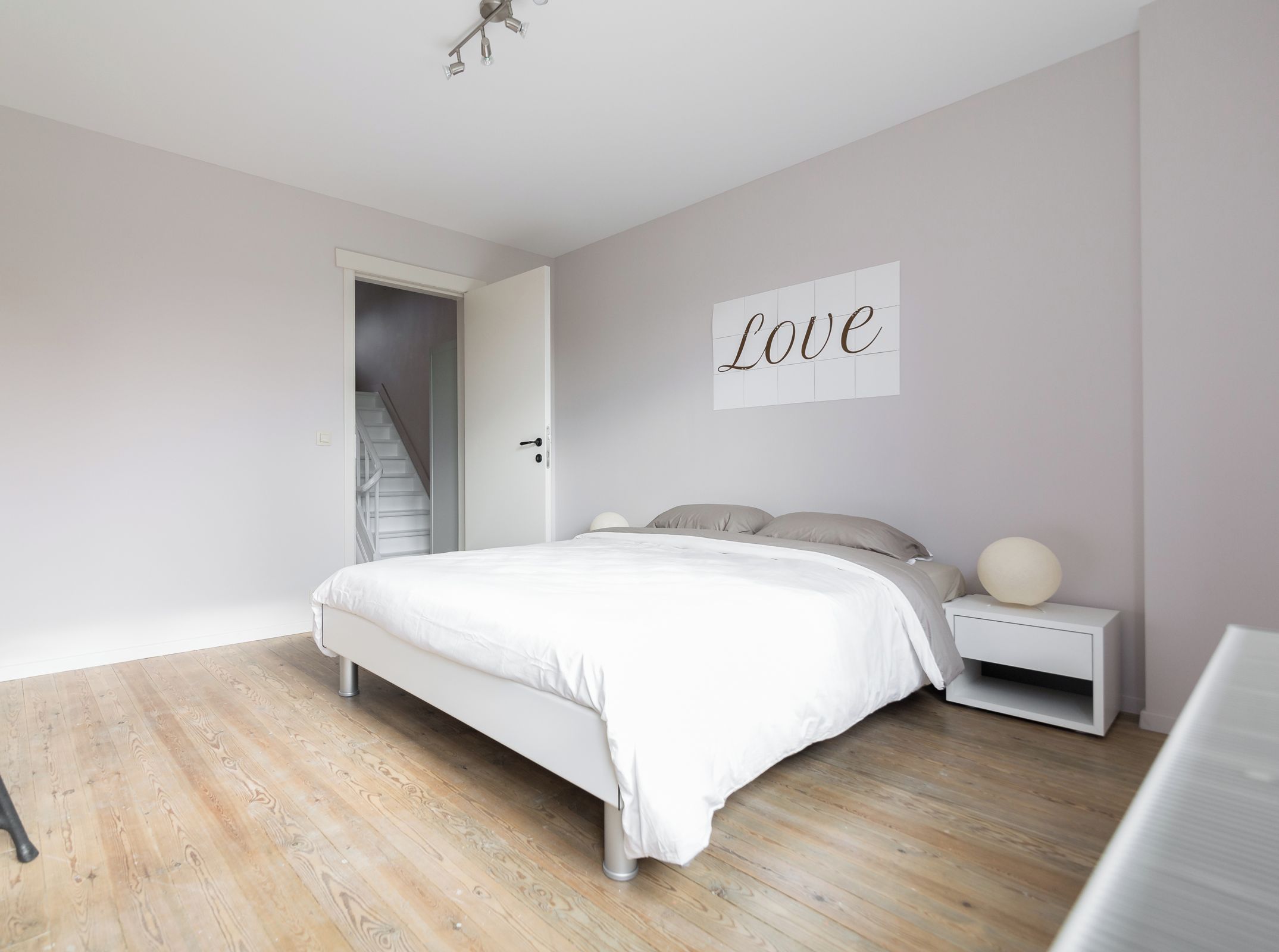 A bedroom with a bed, a nightstand, a wall-mounted sign, and a staircase leading to another room.