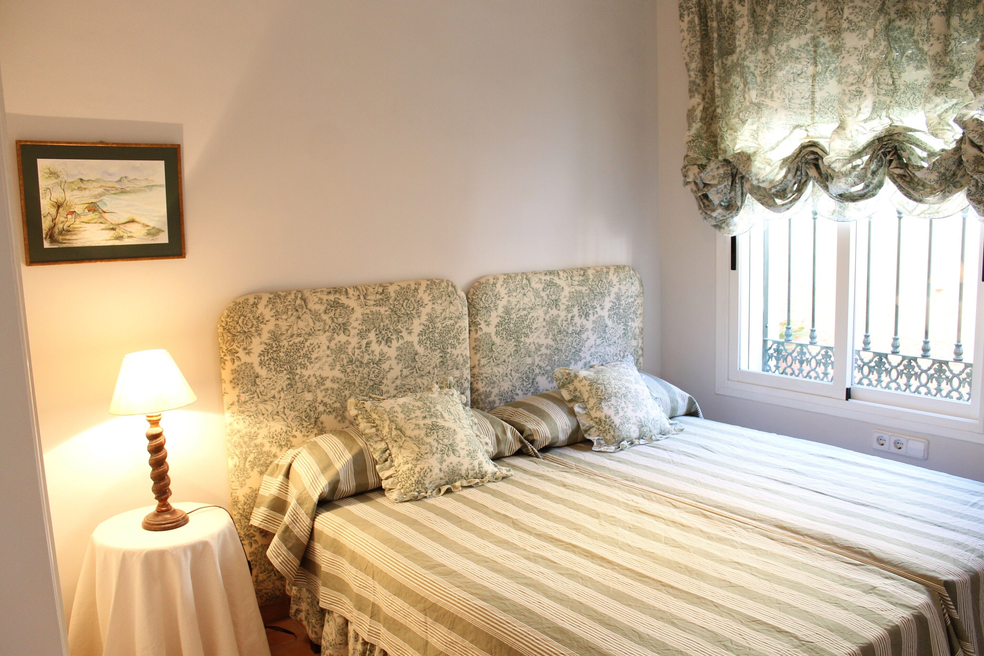A bedroom with a bed, a bedside table with a lamp, patterned pillows, and a window with a view.