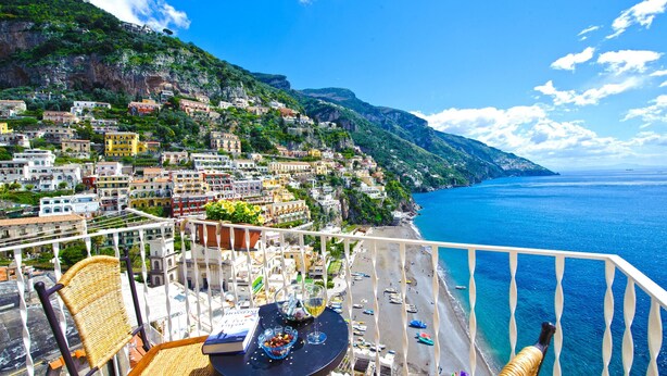 A balcony with wicker chairs, a table with a book and glasses, and a view of a coastal town with buildings and the sea. - Casa Leonilde 4, Emma Villas (Positano)