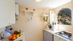 A kitchen with white cabinets, a dishwasher, a stove, and a window with a view of a hillside town. - Casa Leonilde 4, Emma Villas (Positano)