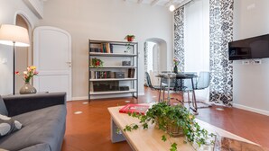 A living room with a sofa, a coffee table with a book, a bookshelf, a dining table with chairs, and a TV mounted on the wall. - Grotta Pinta 6 | Spacious and elegant in the heart of Rome (Roma)