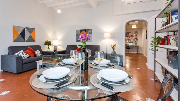 A modern living room with a glass dining table set for four, a grey sofa with colorful cushions, and a bookshelf filled with books and decorative items. - Grotta Pinta 6 | Spacious and elegant in the heart of Rome (Roma)