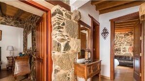 Corredor de paredes de pedra com móveis de madeira e uma cama visível através de uma porta aberta.