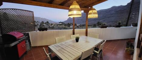Un patio al aire libre con una mesa y sillas de madera, una parrilla y plantas en macetas.