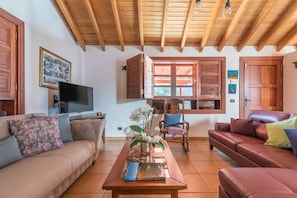 A living room with a wooden ceiling, a television, a sofa, a coffee table, and a window with wooden shutters.