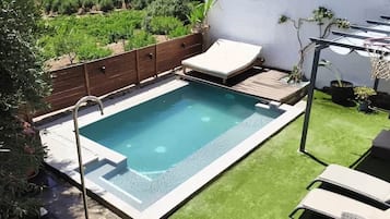 Piscina rectangular con cubierta de madera y sillas de descanso, rodeada por un cerco de madera y follaje.