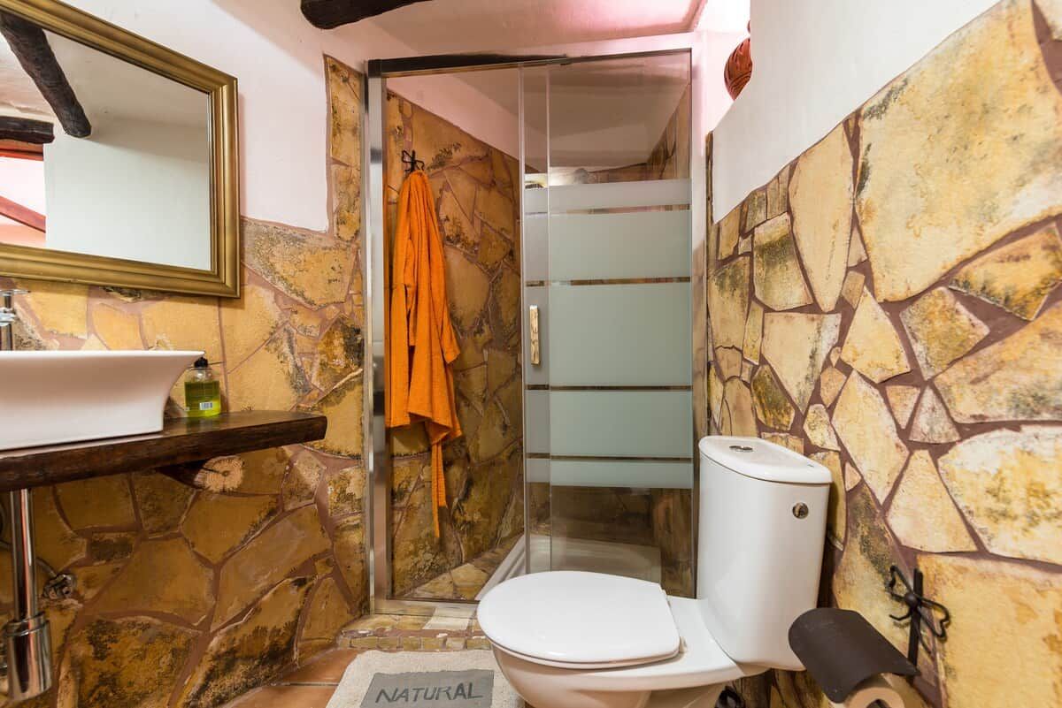 Een badkamer met een stenen muur, een wit toilet, een douchecabine met glazen wanden en een spiegel.
