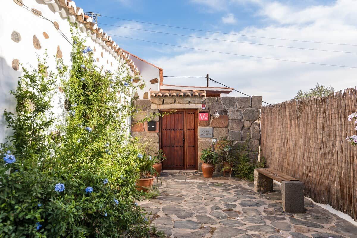 Un sendero de piedra que lleva a una puerta de madera, rodeado de macetas con plantas y un muro de piedra.