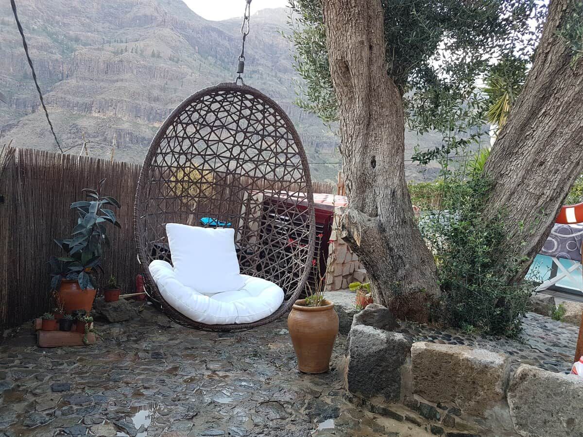 Una silla colgante de mimbre con un cojín blanco, una cesta colgante y plantas en macetas en un patio con montañas al fondo.