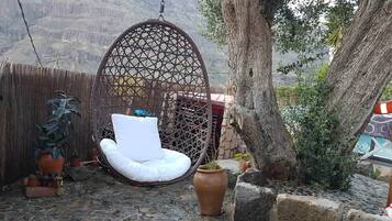 Una silla colgante de mimbre con un cojín blanco, una cesta colgante y plantas en macetas en un patio con montañas al fondo.
