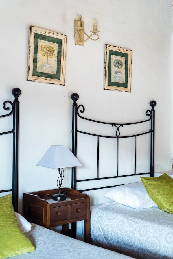 A bedroom with a metal bed frame, a bedside table with a lamp, two framed pictures on the wall, and a green pillow on the bed.