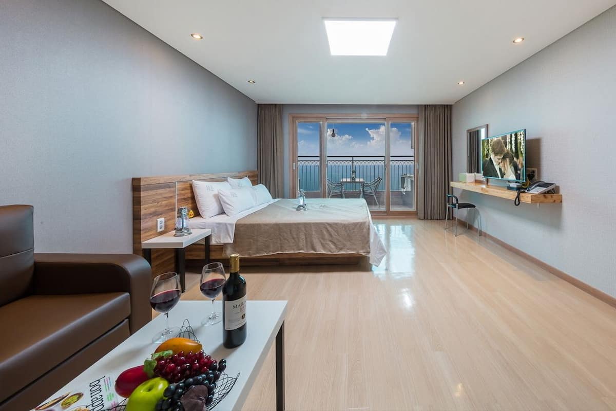 A hotel room with a bed, a TV, a balcony with a table and chairs, a dining table with fruits and wine, and a sofa.