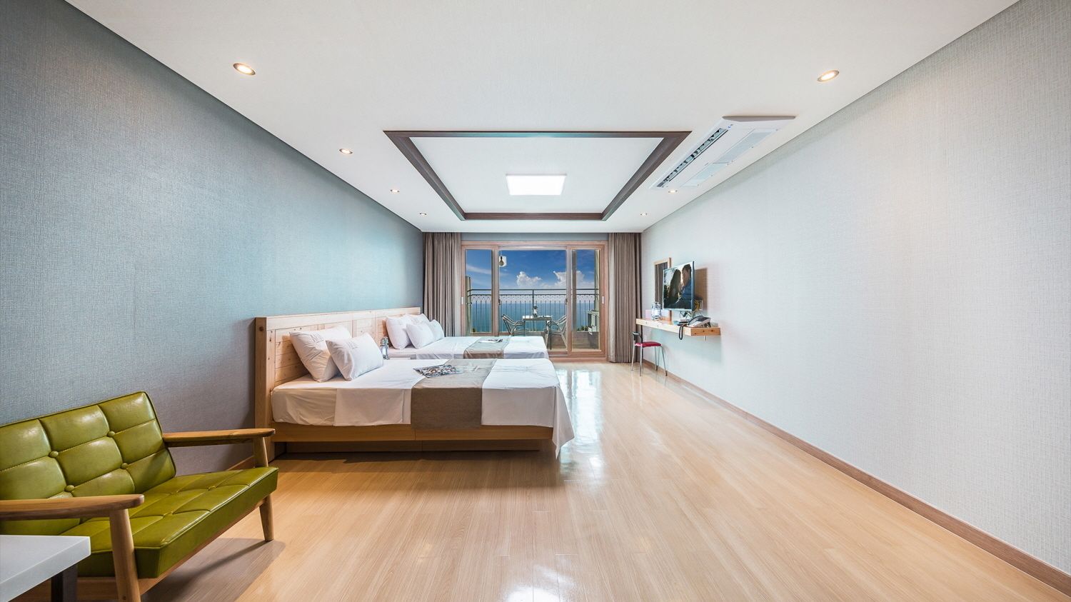 Una habitación de hotel moderna con una cama grande, un sofá verde, un escritorio con una silla y vistas a la ciudad a través de amplios ventanales.