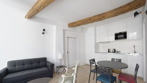 A modern kitchen with white cabinets and a microwave. There is a dining area with a round table and chairs, and a grey sofa in the living area.