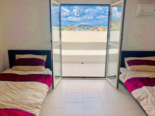 A room with two beds, a sliding glass door leading to an outdoor area, and a view of a building with a green roof.