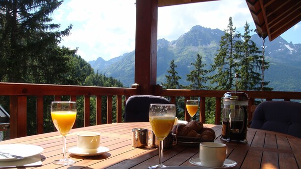A wooden table set for breakfast with croissants, juice glasses, and a coffee pot, overlooking a mountainous landscape.
