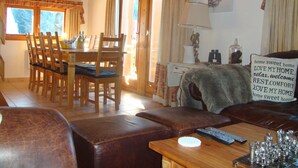 A cozy living room with a wooden dining table, chairs, a sofa, and a coffee table. There is a lamp, a sign with a message, and a deer painting on the wall.