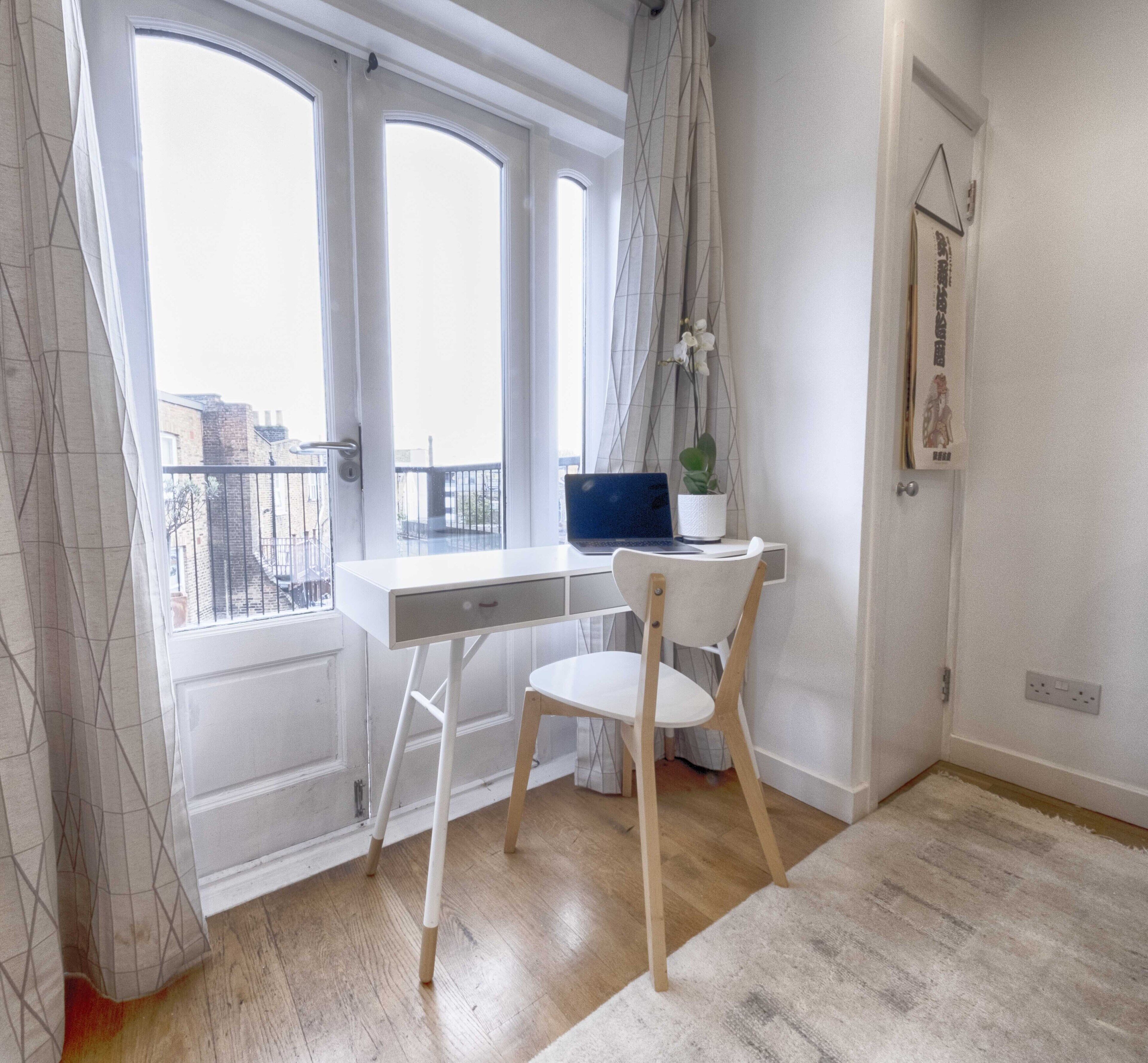 Ein Zimmer mit Schreibtisch, Stuhl, Laptop und einem Fenster mit Vorhängen.