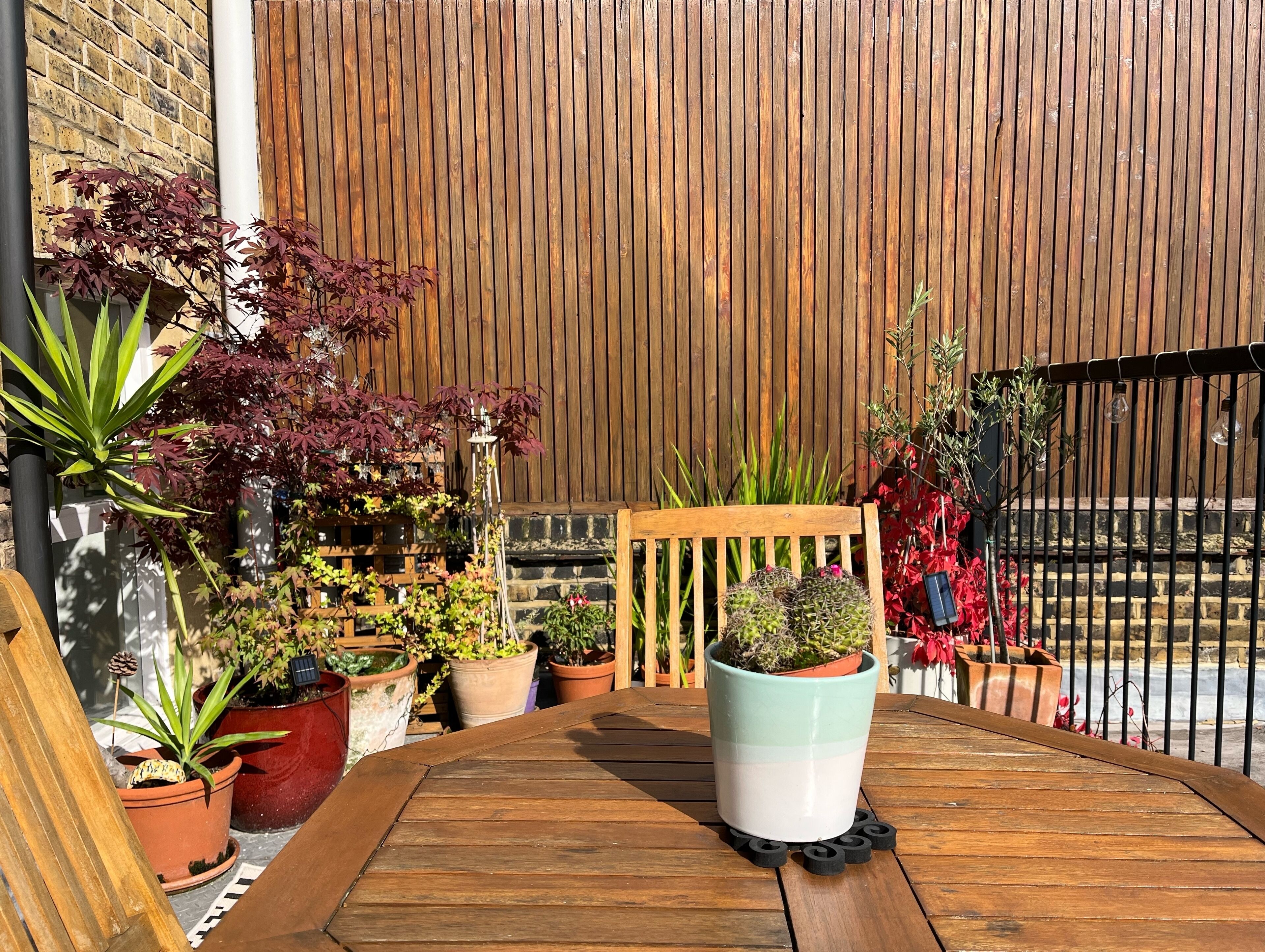 A wooden table with potted plants and a bench in a garden area with a wooden fence and brick wall in the background.