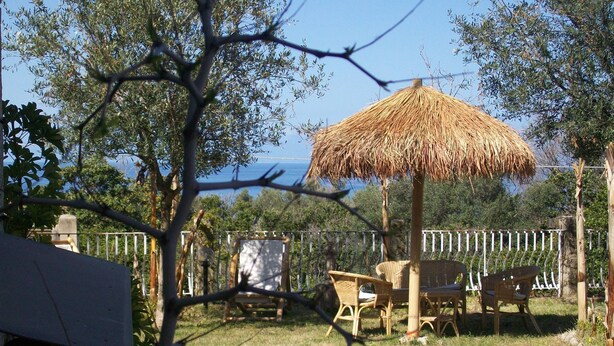 Un área de estar al aire libre con mobiliario de mimbre y un toldo de paja, rodeada de árboles y vistas al mar.